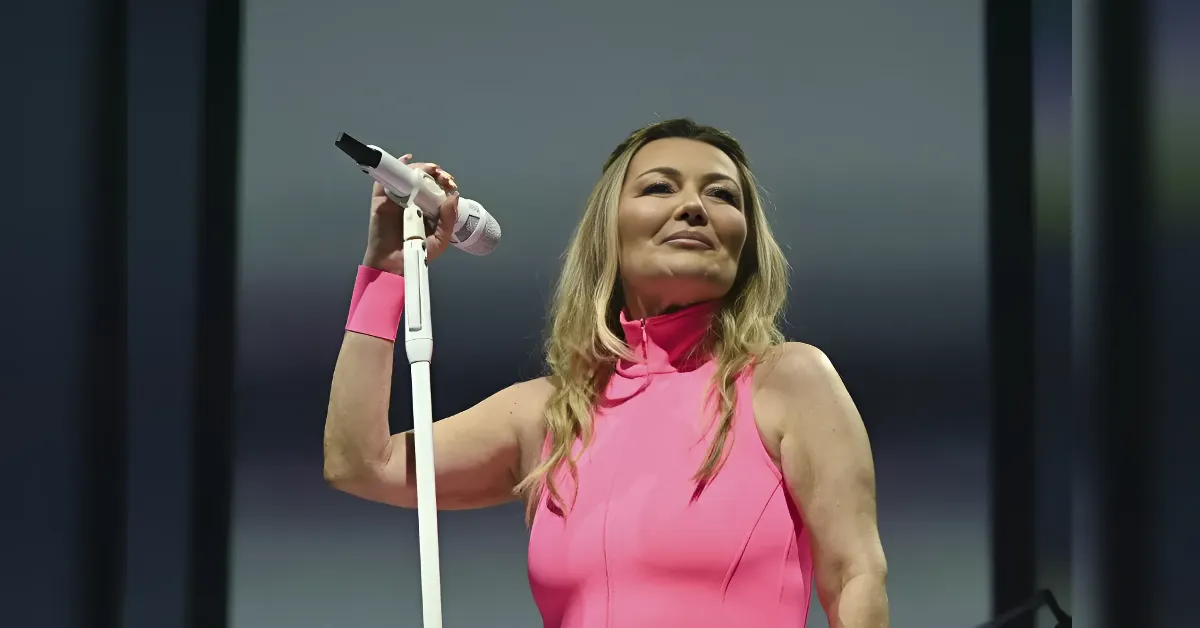 Female singer in a pink sleeveless top performs on stage, gripping a white microphone stand with a pink wristband visible on her arm.