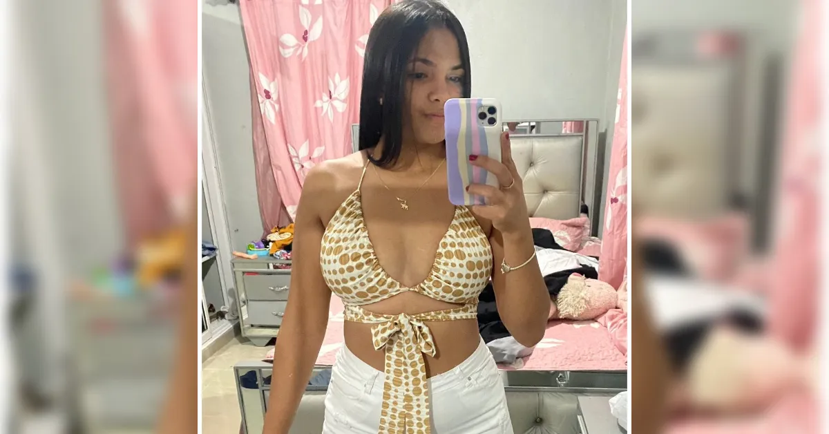 Woman taking a mirror selfie in a pink-themed bedroom, wearing a beige polka-dot bikini top and white pants.