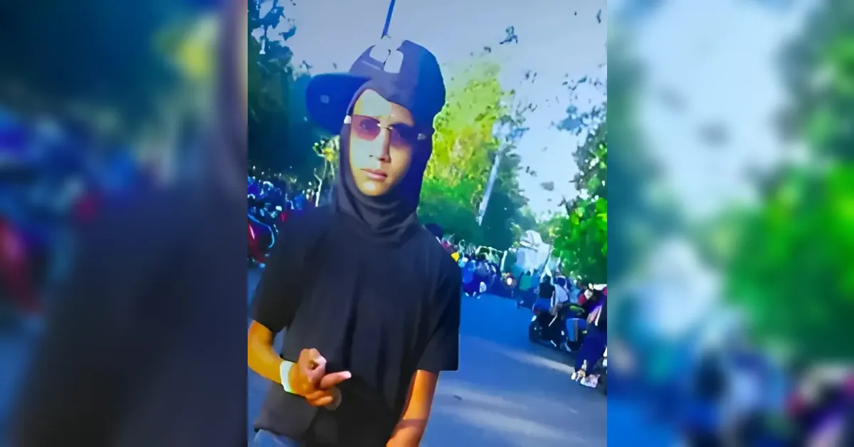 Person in a black hoodie and hijab with a cap and sunglasses stands in a street with spectators watching a parade; the person gestures with a raised hand.