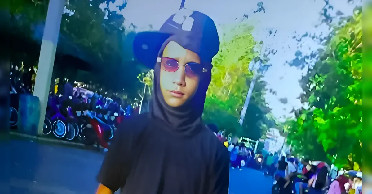 Person in a black hijab and cap with sunglasses stands on a sunny street, crowd seated along the path in the background.