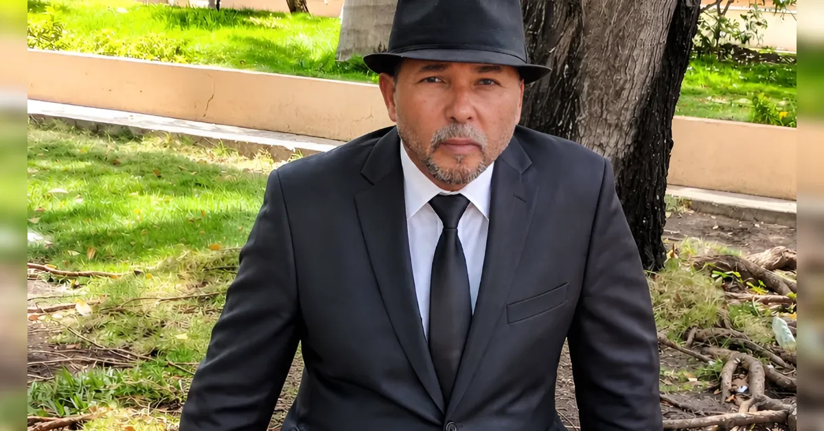 Man in a black suit and tie wearing a fedora, sitting on the grass near a tree in a park setting.