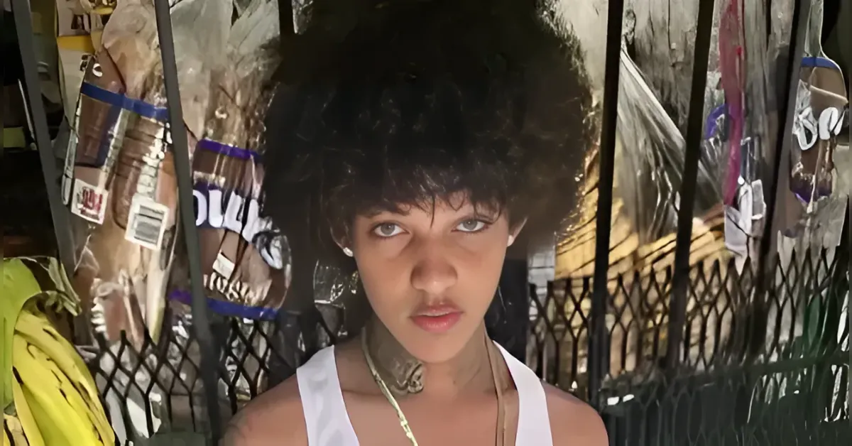 Close-up portrait of a young person with short black hair and a white tank top, looking intently at the camera behind a wrought-iron fence with posters attached to it.