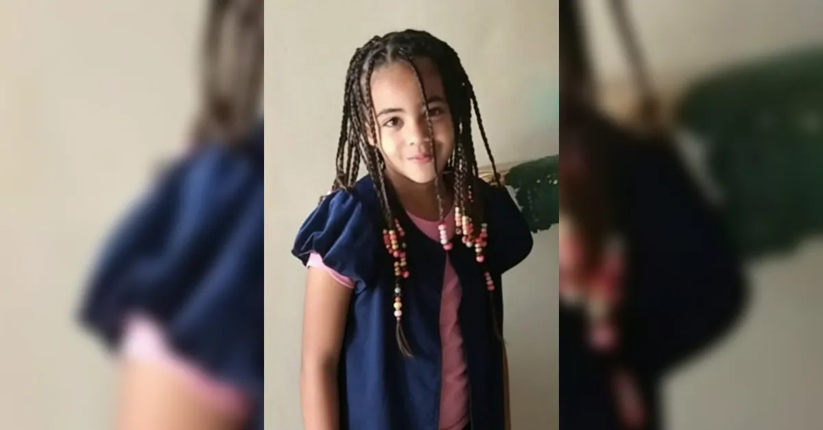 Young girl with braided hair wearing a navy dress over a pink shirt, smiling at the camera.