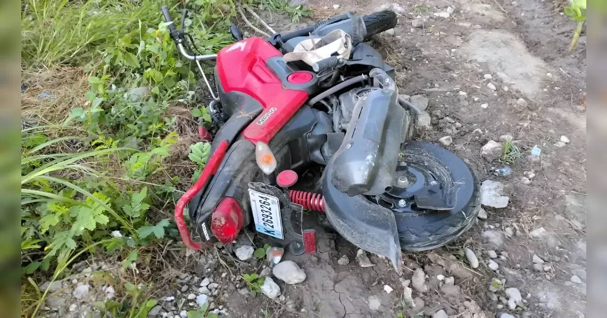 Red scooter lying on its side on a muddy dirt surface, license plate visible behind the rear wheel and damaged body parts nearby.