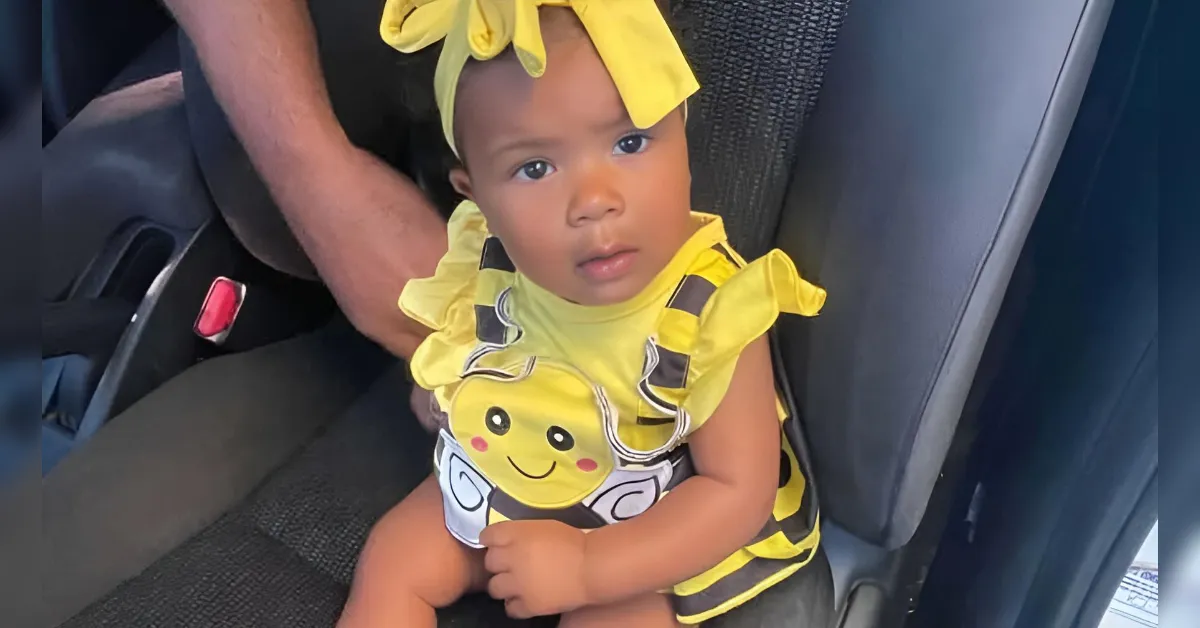 Toddler in a yellow bee costume with a bow headband sitting in a car seat, looking at the camera