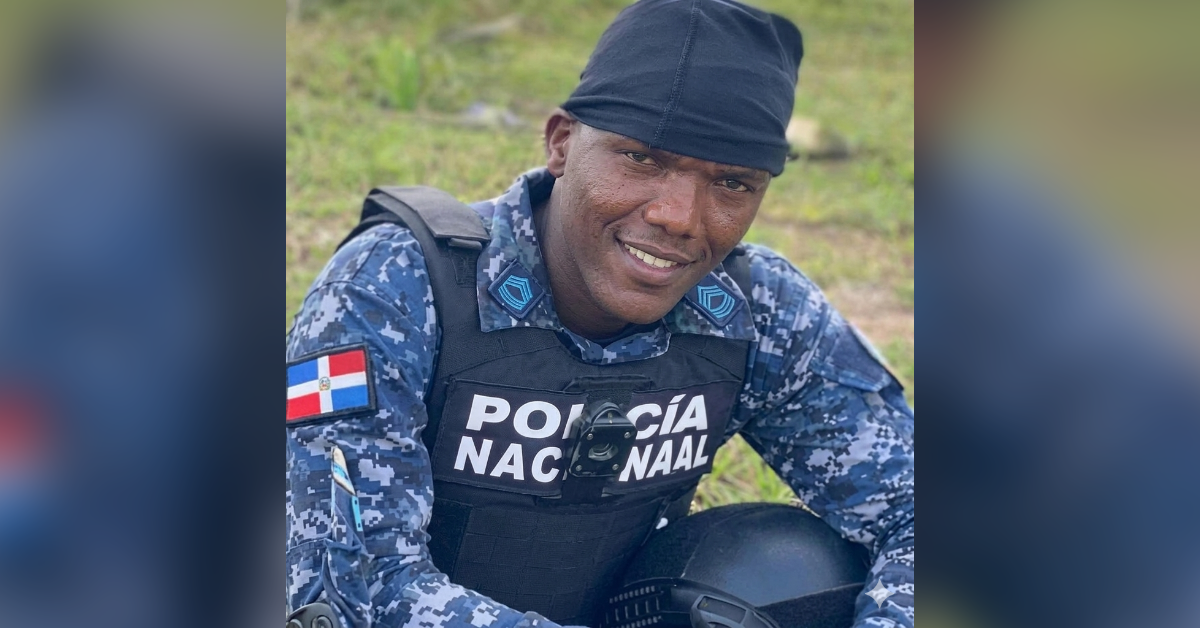 Dominican Republic police officer in camouflage uniform smiling outdoors wearing a tactical vest with Policía Nacional text and Dominican flag patch on the sleeve.