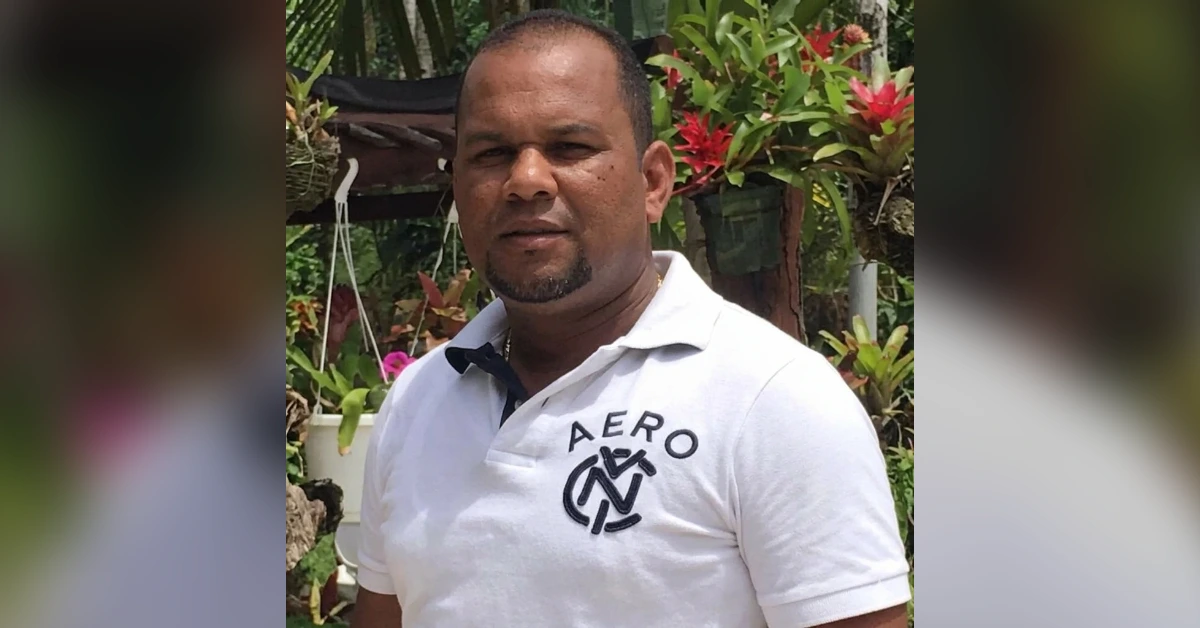 Man wearing a white polo shirt with an Aero logo stands in a lush garden with green plants and red flowers in the background, facing the camera.