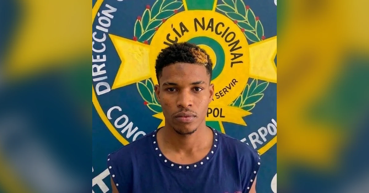 Young man wearing a navy blue shirt with studded neckline, standing in front of a large police insignia backdrop.