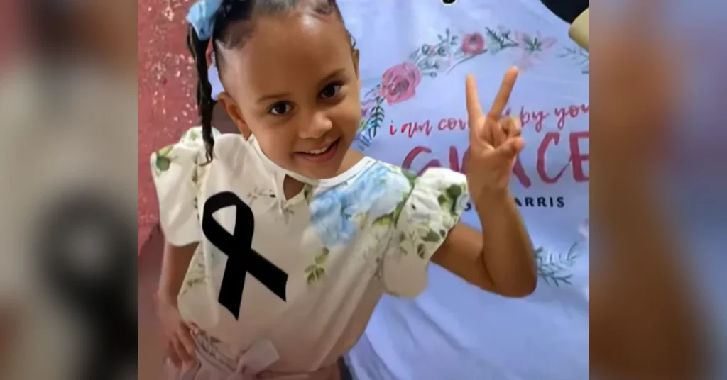 Young girl smiles and shows a peace sign with two fingers, wearing a white floral shirt with a black cancer-awareness ribbon pinned to it, in front of a banner.