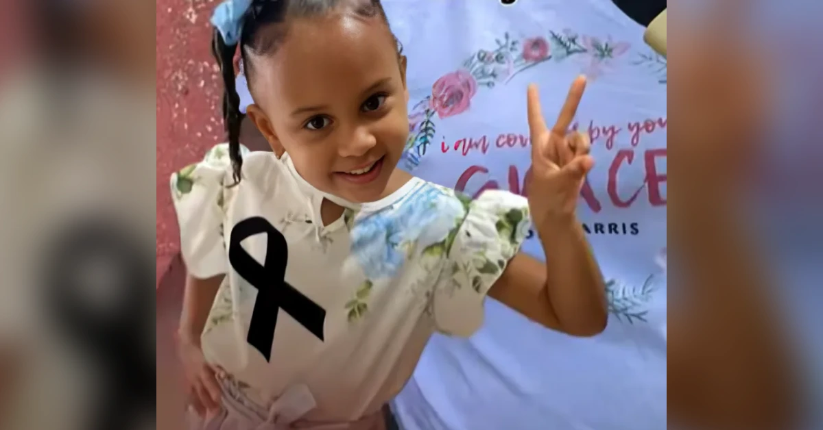 Smiling young girl in a floral dress shows a peace sign with her right hand, wearing a black cancer-awareness ribbon on her chest, standing in front of a banner with pink flowers.