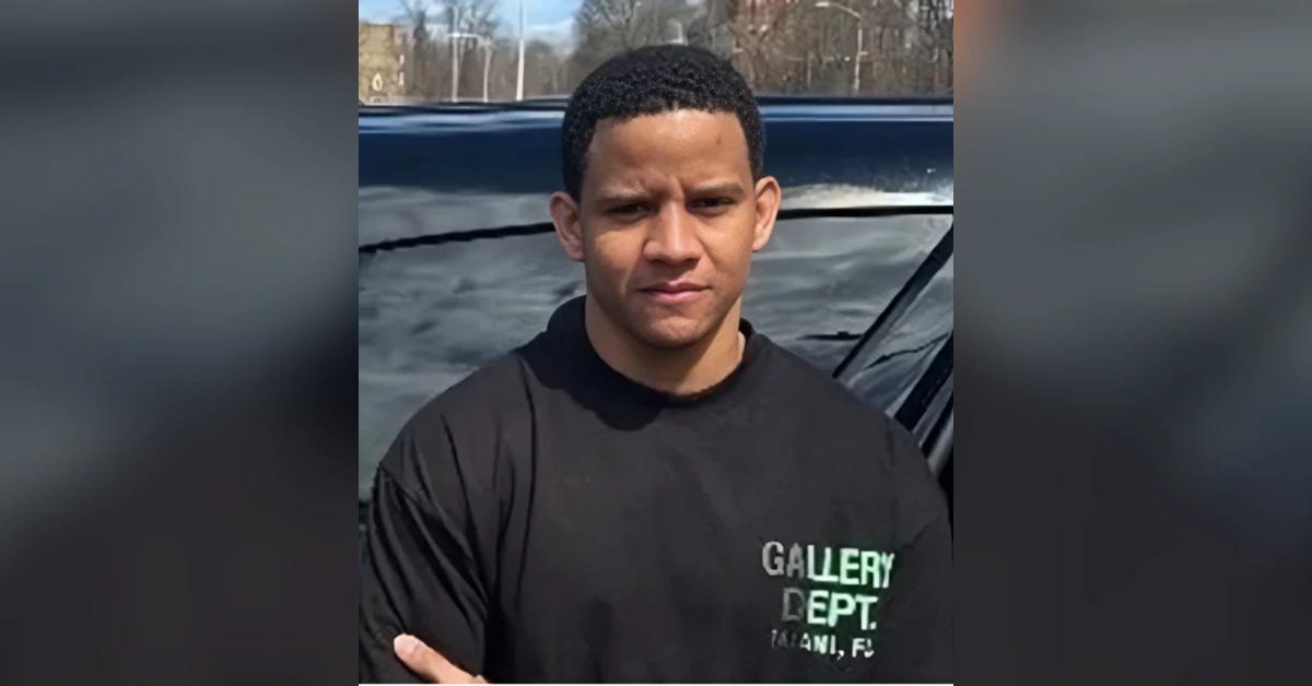 Young man with short curly hair in a black Gallery Dept. T-shirt stands in front of a dark car outdoors.