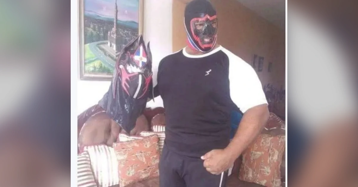 Muscular person in a black-and-white athletic outfit wearing a red wrestling mask stands indoors holding a decorative lucha libre mask.