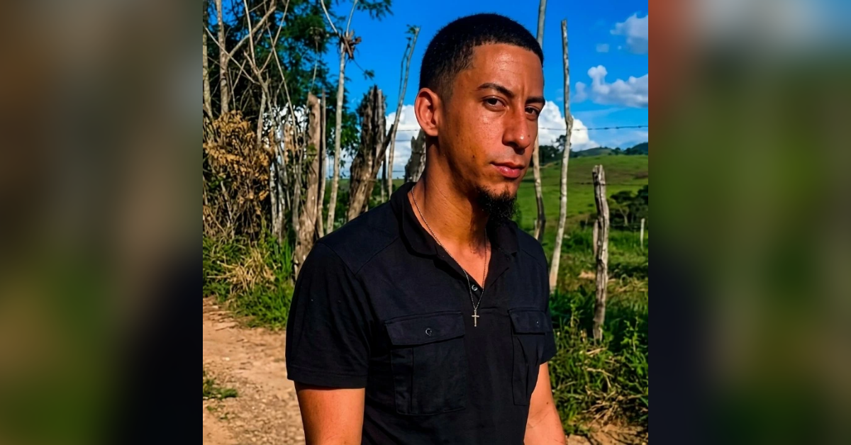 Portrait of a man with short hair and goatee, wearing a black polo with a cross necklace, standing on a dirt path with wooden fence posts and green hills in the background under a blue sky.
