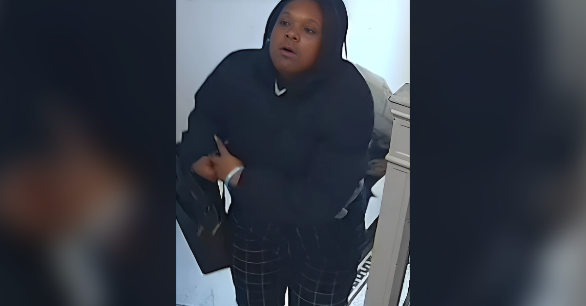 Woman in a black hooded sweatshirt and plaid pants stands indoors, looking slightly upward.