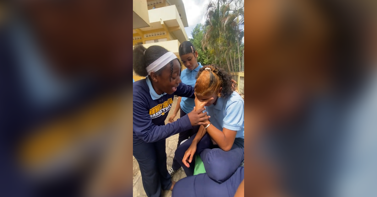 Three students in blue school uniforms comfort a crying classmate outdoors in a campus courtyard.