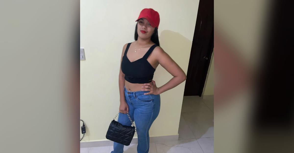 Woman in a red cap, black crop top, and blue jeans posing indoors with a black quilted handbag.