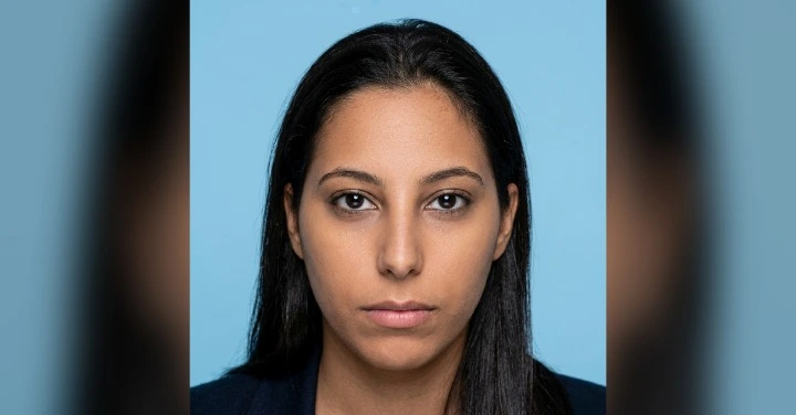 Portrait of a woman with dark hair and a neutral expression facing the camera against a light blue background.