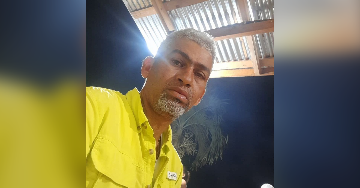 Man with gray beard wearing a bright yellow shirt, taking a selfie at night under a corrugated roof.