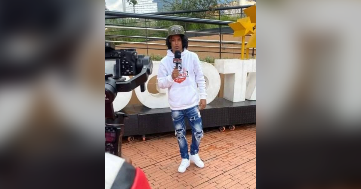 Young man in a white hoodie and ripped jeans speaks into a microphone while a camera films him outdoors.