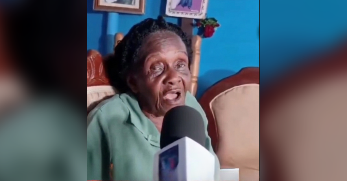 Elderly woman in a green shirt speaks into a microphone during a home interview, with a blue wall in the background.