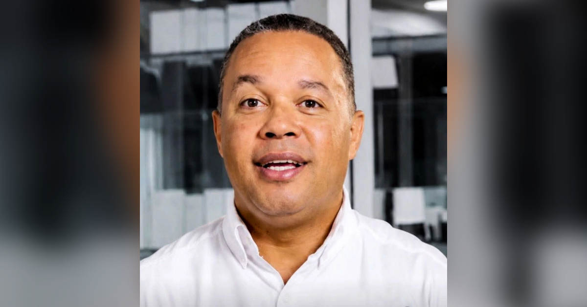 Smiling man in a white shirt, speaking, with an office backdrop behind him.