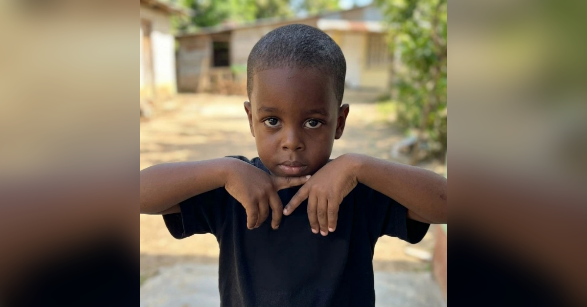 Boy with short hair wearing a black shirt, arms crossed in front of him, looking at the camera outdoors.