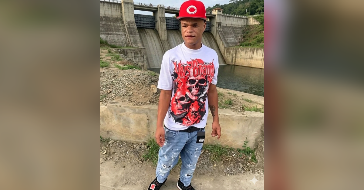Young man standing outdoors near a dam, wearing a red cap, white graphic tee, and distressed jeans.