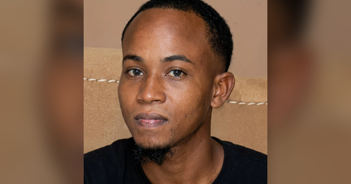 Close-up portrait of a man with a short black beard and mustache, wearing a black shirt against a beige background.