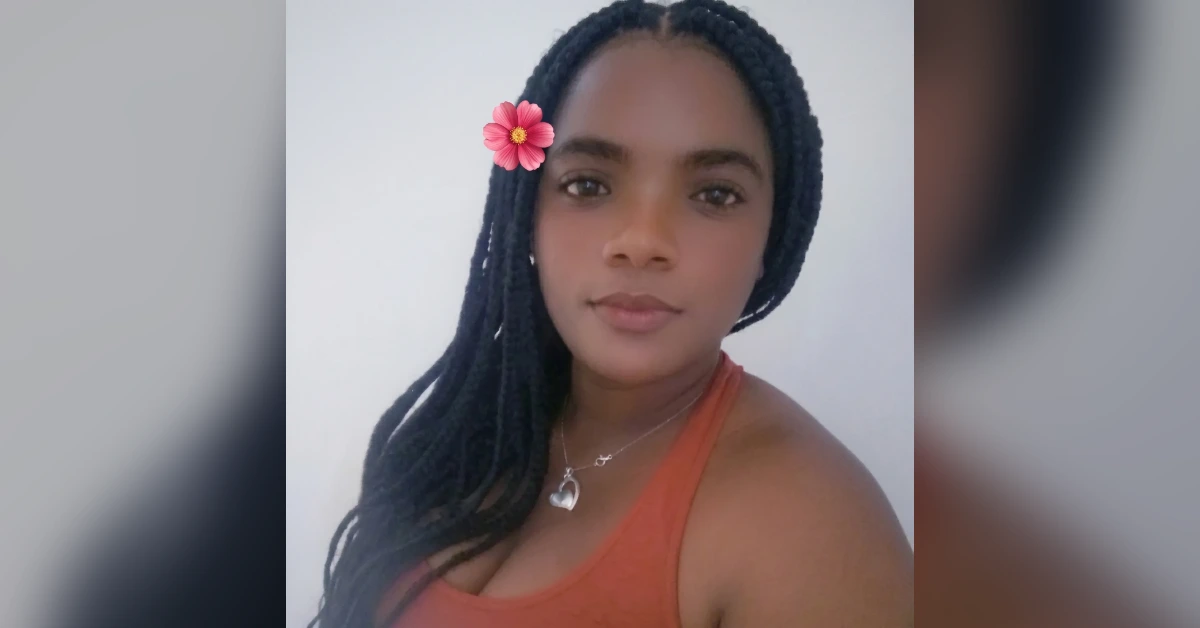 Portrait of a woman with braided hair, a pink flower in her hair, an orange sleeveless top, and a heart necklace, looking at the camera against a light background.