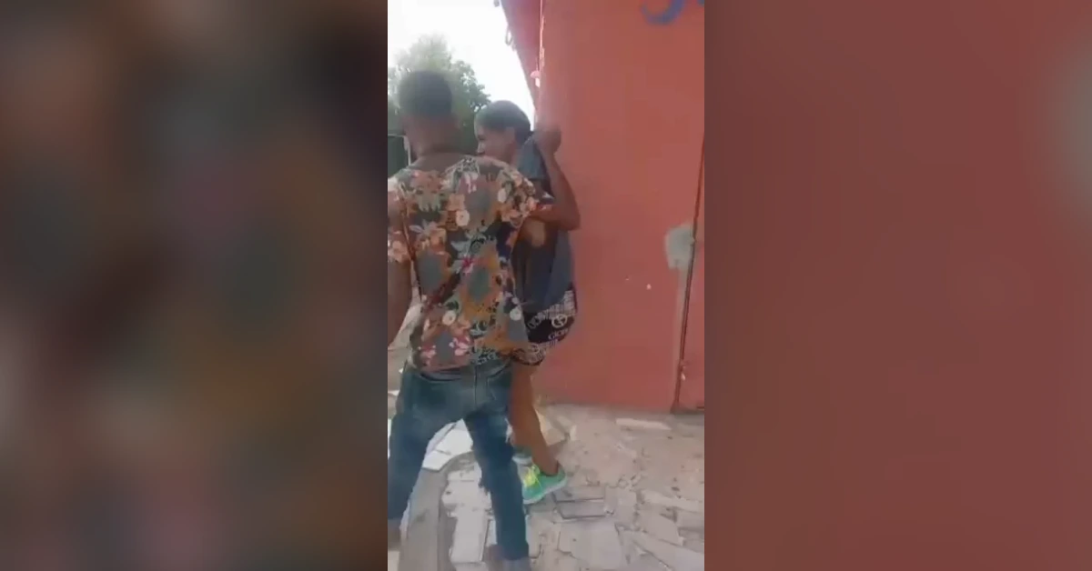 Two teens on a sidewalk by a red wall. One in a bright floral shirt grabs the other by the arm, as they stand close together on cracked pavement.