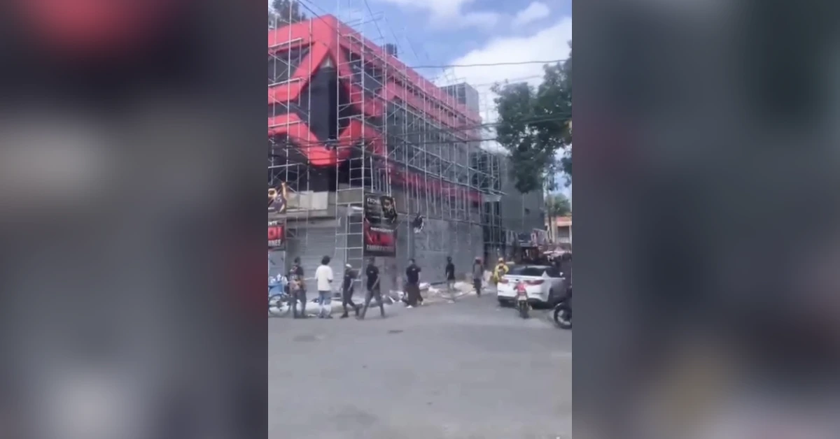 A city street with a building under construction wrapped in red scaffolding; pedestrians and vehicles pass by on a sunny day.