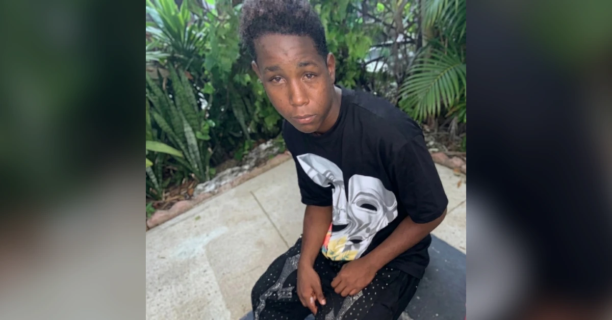 Young man sits outdoors on a concrete surface, wearing a black graphic t-shirt and dark shorts, gazing at the camera with a neutral expression.