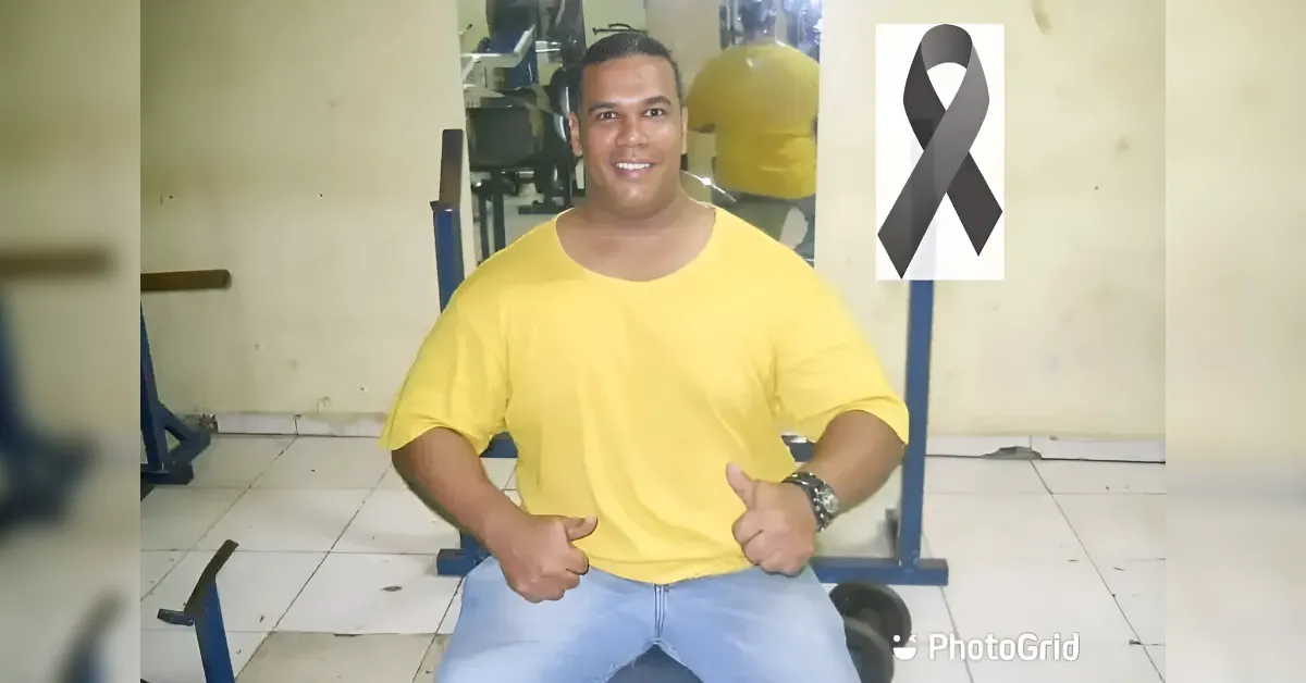 Smiling man in a yellow shirt sits on a gym bench and gives thumbs up, with gym equipment in the background and a black awareness ribbon symbol nearby.