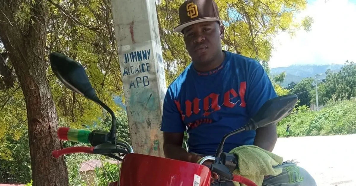 Man wearing a brown cap and blue shirt sits on a red motorcycle outdoors beside a graffiti-covered pole.