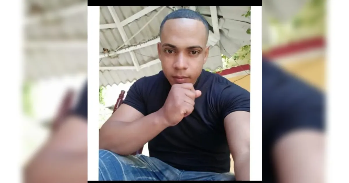 Young man in a black t-shirt sits outdoors, resting his chin on his fist and looking at the camera.