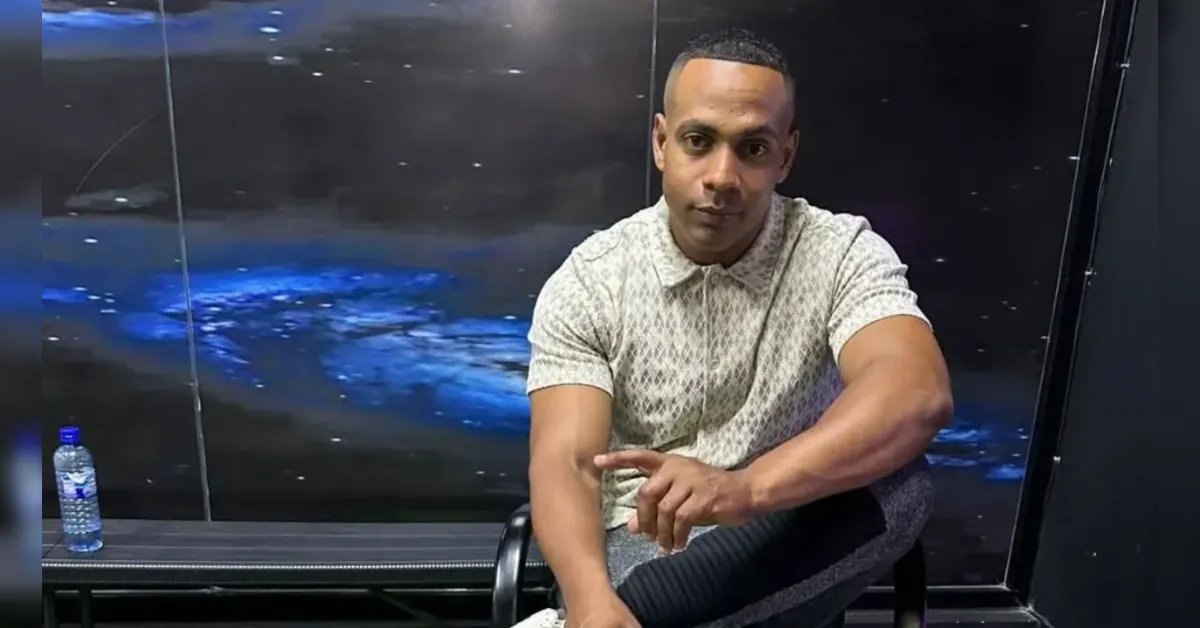 Man seated on a chair in a dark studio with a glass wall and a blue-lit background, wearing a light patterned polo shirt and dark joggers. A water bottle sits on a table nearby.