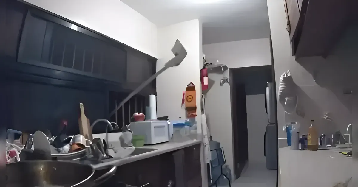 Cluttered galley-style kitchen with a sink and dishes on the left counter, a microwave, and various containers; a refrigerator is on the right side in a narrow room with dim lighting.