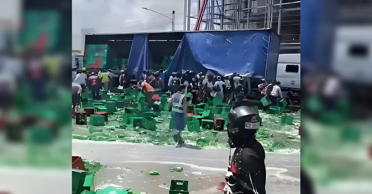 Truck with blue tarpaulin unloading green plastic crates onto a street as people gather amid scattered boxes and green liquid on the ground in a busy loading area.