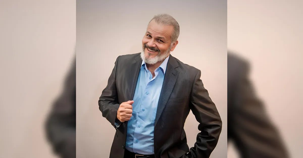 Smiling man in a dark suit and blue shirt with one hand on his jacket lapel, posing confidently against a neutral background.
