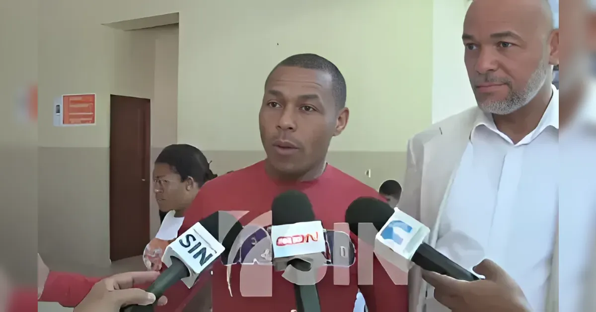 Man in a red shirt speaks to reporters holding microphones in a hallway.