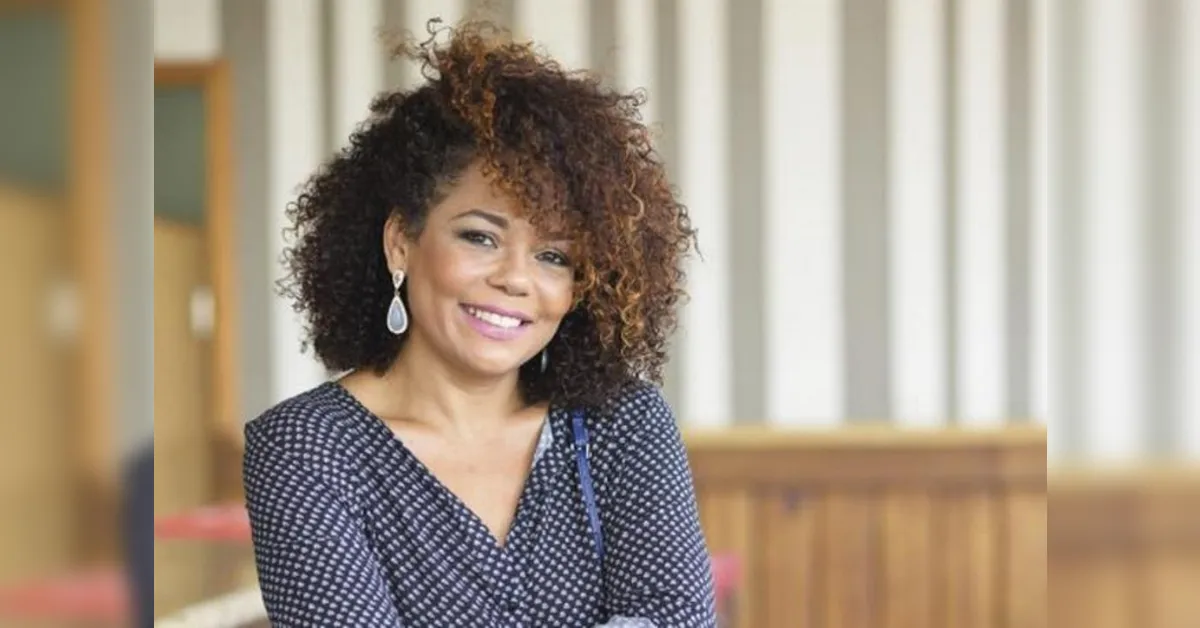 Smiling woman with curly hair wearing a dotted blouse, posed indoors with a bright background.