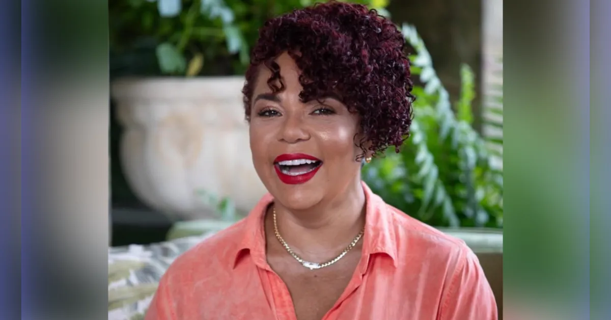 Smiling woman with burgundy curly hair in a coral shirt, wearing a necklace, outdoors with greenery behind her.