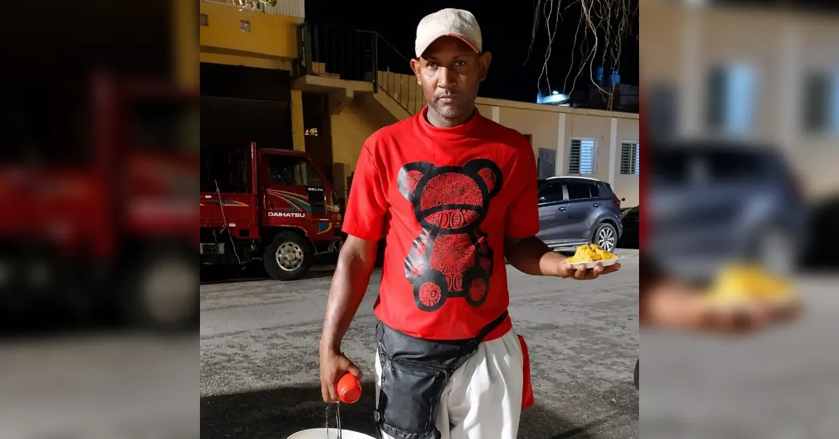 Man in a red t-shirt and white cap stands on a street at night, holding a plate of yellow rice and a red bottle/condiment in his other hand, with a bag around his waist.