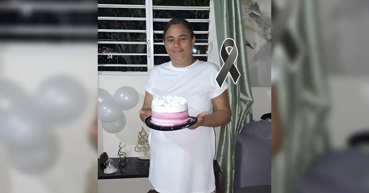 Person in a white shirt holding a pink-and-white frosted cake on a plate indoors, with balloons in the background.