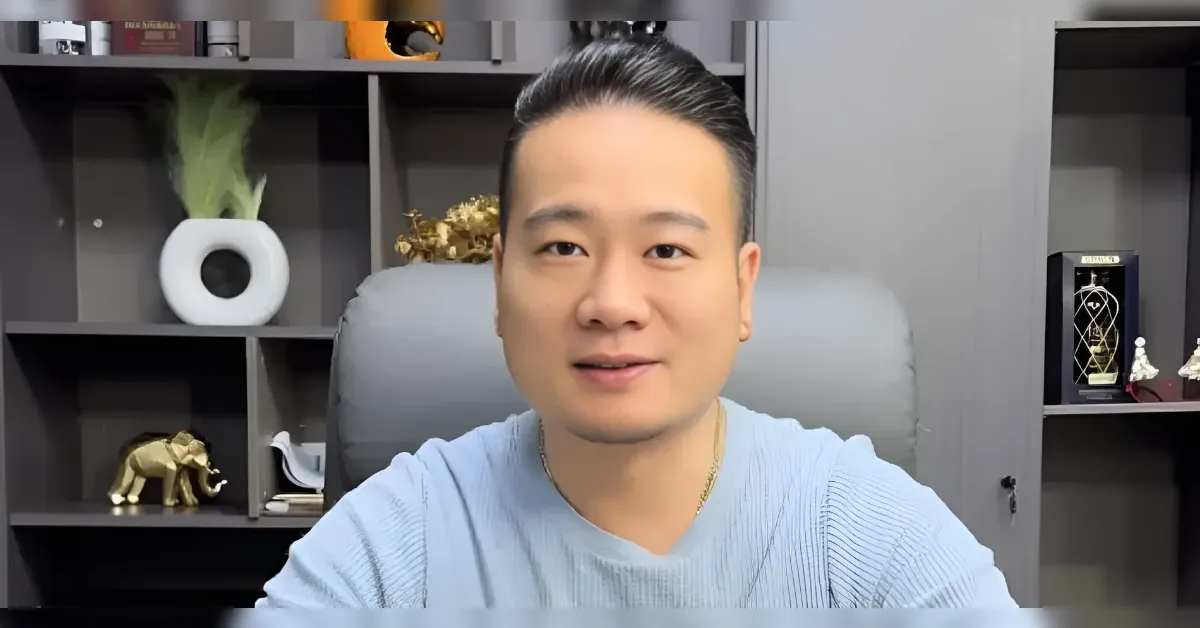 Man with dark hair tied back sits in a gray office, wearing a light blue sweater, looking at the camera with shelves and decorative items behind.