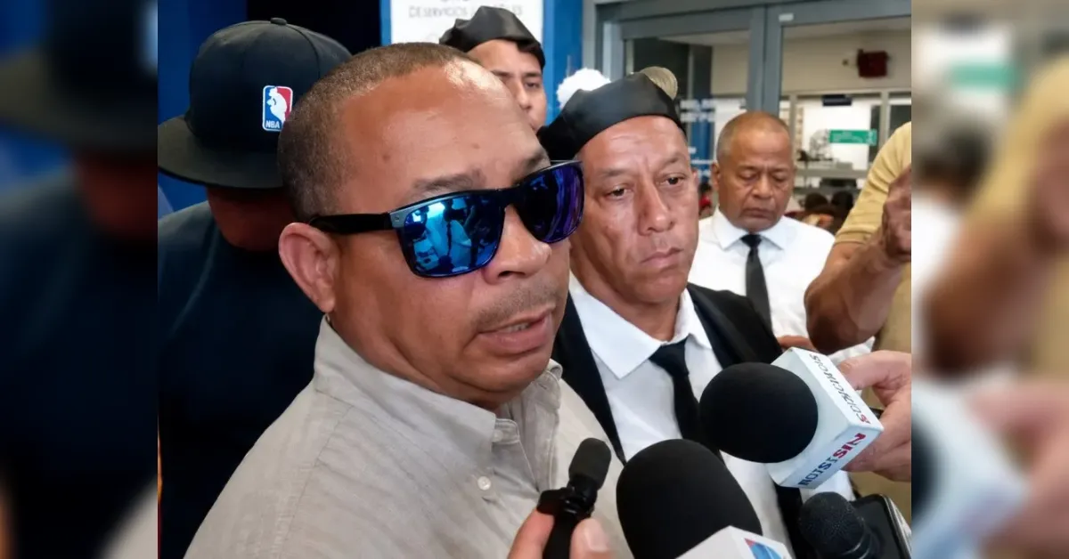 Man in beige shirt and blue sunglasses is interviewed by multiple reporters, microphones close to his face in a crowded press area.