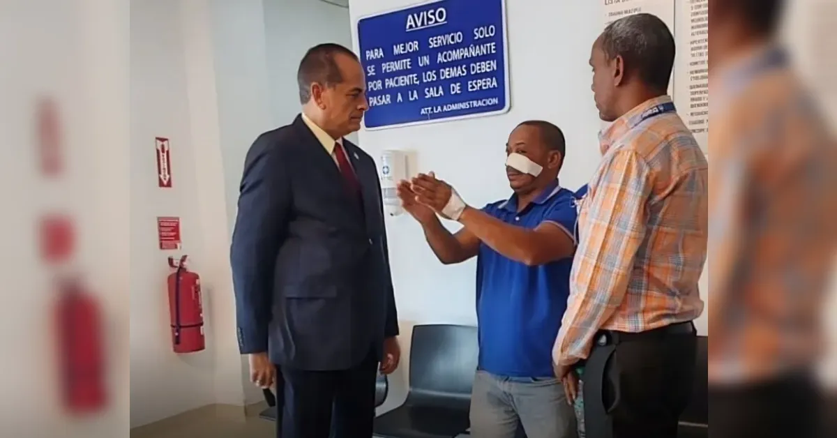 Man in a dark suit speaks to a blindfolded man in a blue polo while a third man in a checkered shirt looks on in a hallway; fire extinguisher and a blue AVISO sign are in the background.