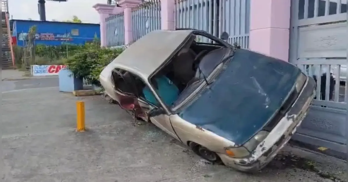 Beige sedan tipped onto its left side against a metal gate, front end scraped and windshield cracked, car upholstery visible through the open window. Parking bollard and urban surroundings in the background.