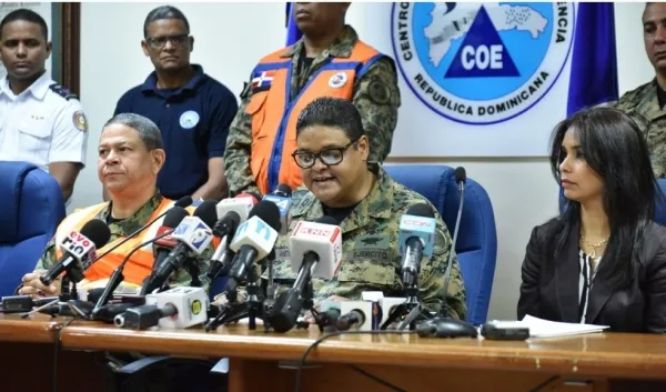 Colonel speaking at a Dominican civil defense press briefing, with numerous microphones in front and colleagues, COE banner in the background.