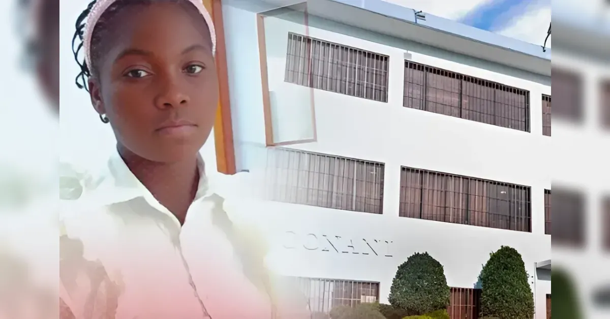 Portrait of a young person in a white shirt and headband standing in front of a white, multi-story building with large barred windows and trimmed bushes.